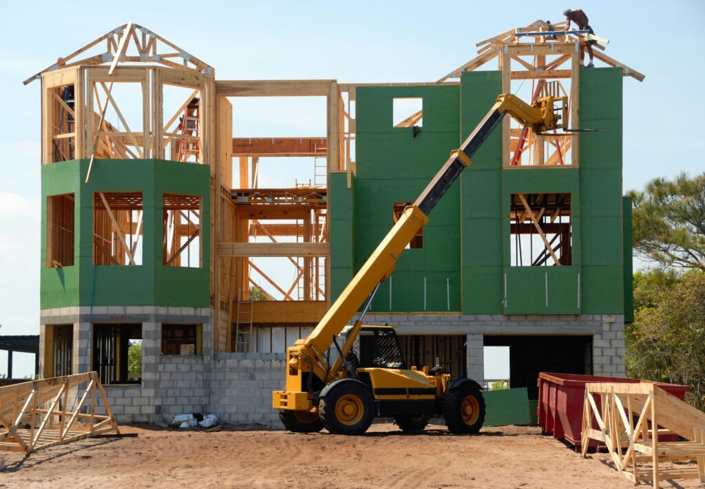 A large house under construction with exposed wood framing, green sheathing panels, concrete block foundation, and a yellow telescopic handler lifting materials to the upper level, with workers on the roof.