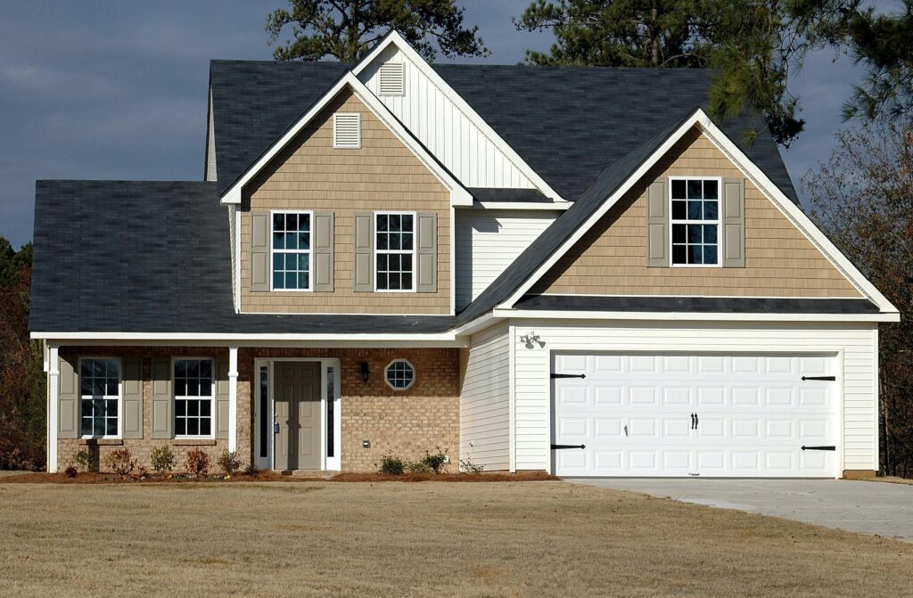 A completed two-story suburban house with beige siding, white trim, and a white double garage door, set on a manicured lawn with trees in the background