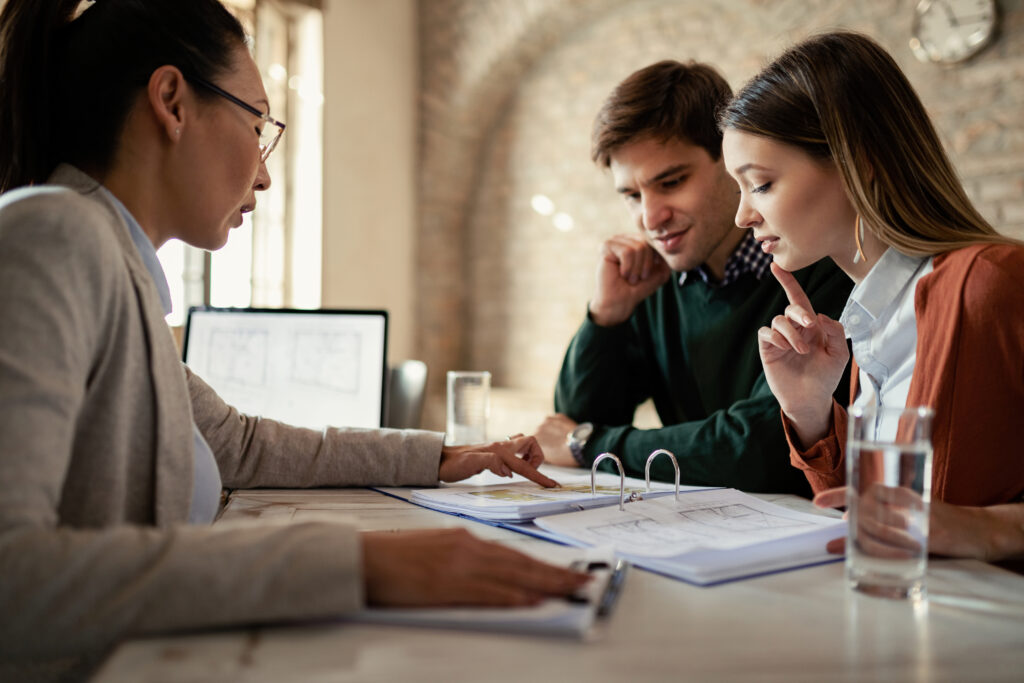 Businesswoman consulting with a couple at a desk, discussing documents with a computer screen visible in the background in a professional office setting.

