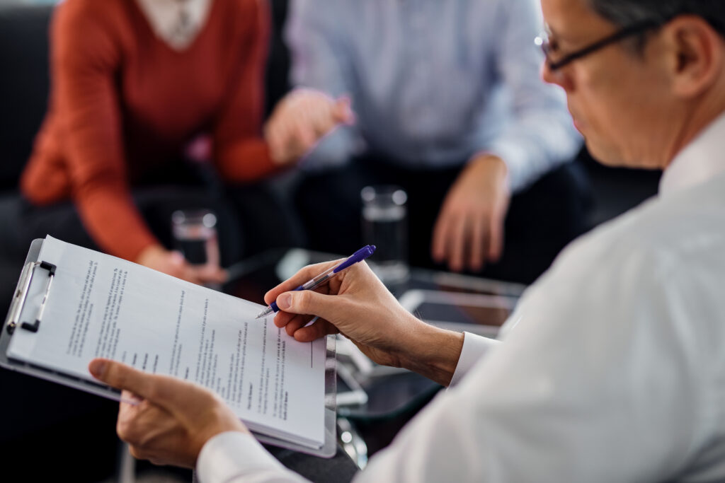 Close-up of an advisor filling out a form on a clipboard while sitting with two clients during an insurance consultation, with glasses of water on the table.