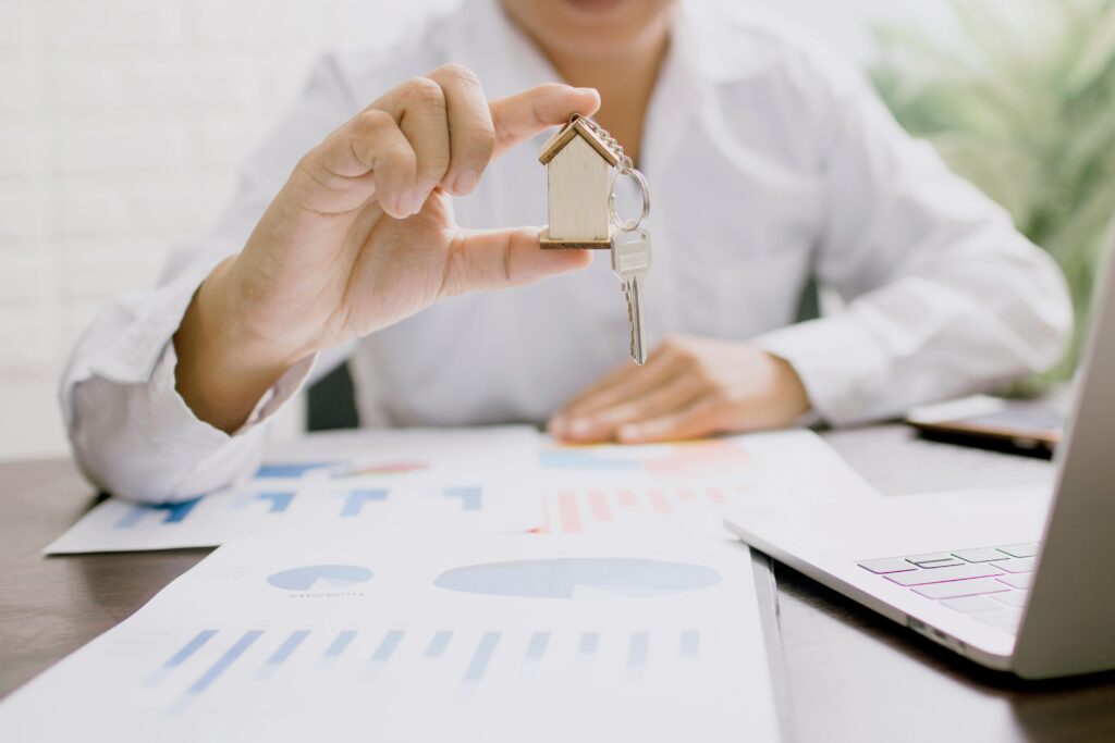 A person in a white shirt holding a small wooden house keychain with a key, sitting at a desk covered with financial charts and a laptop