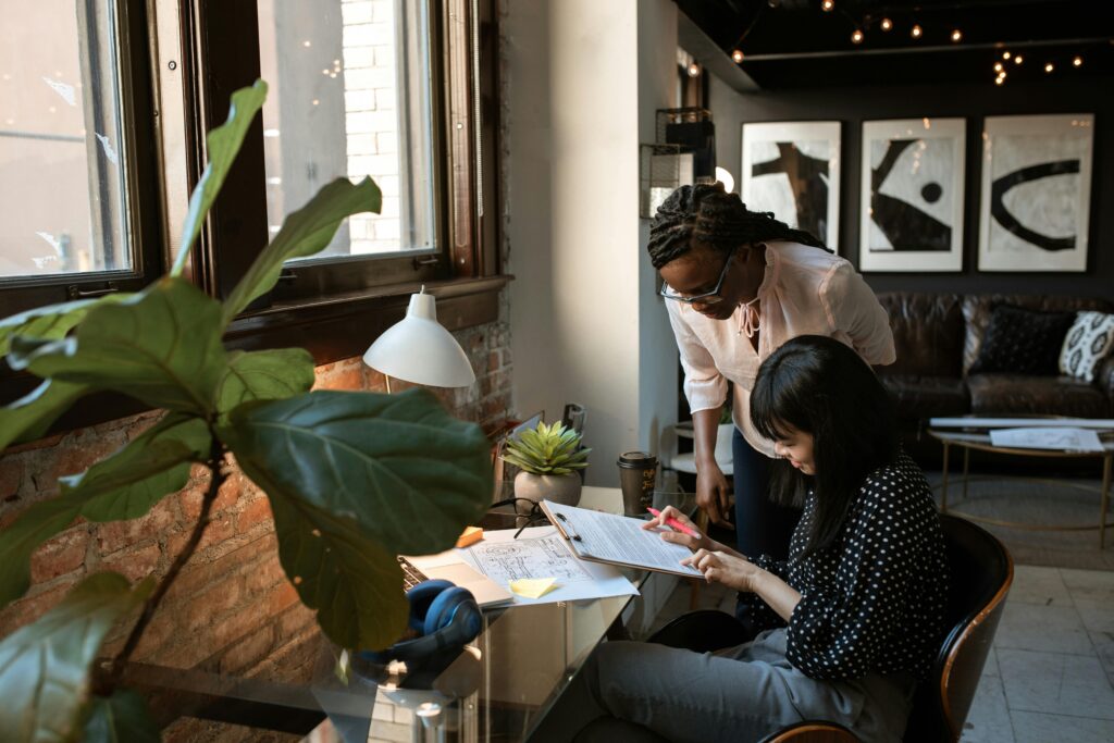 Two women work together at a desk in a modern office. One woman sits and reviews documents with a highlighter, while the other stands beside her, offering guidance. The workspace features a large window, potted plants, and abstract art on the wall in the background.