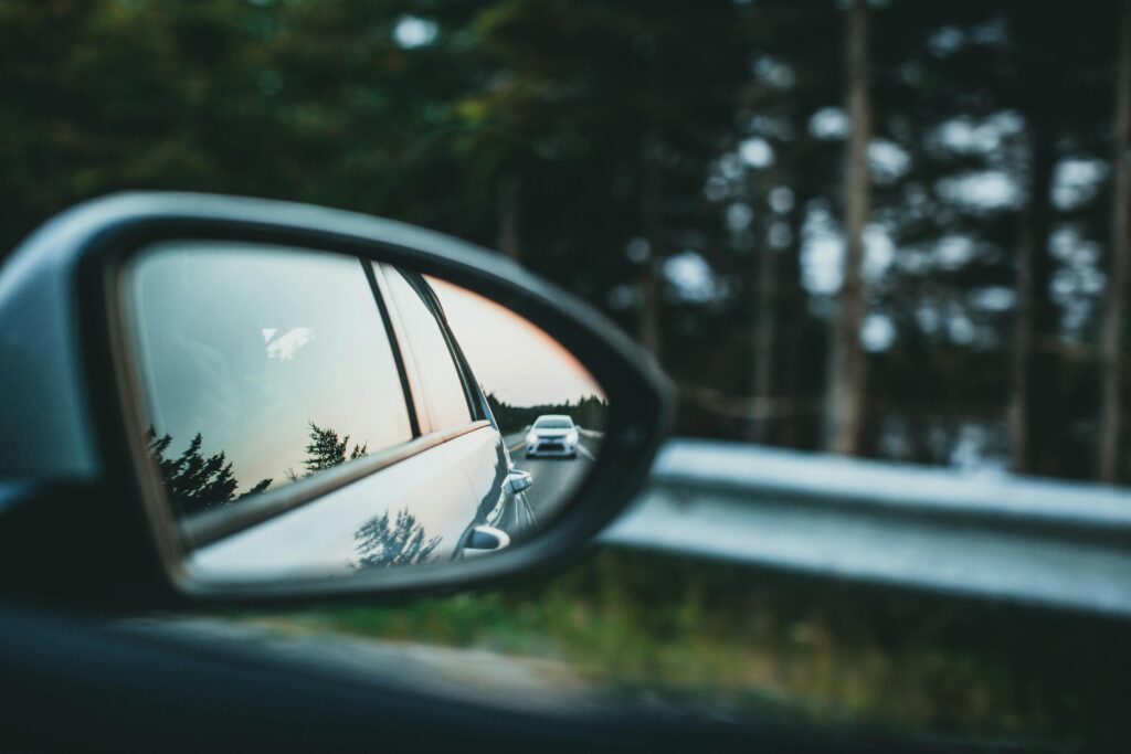 A close-up view of a car’s side mirror reflecting a white vehicle driving on a forest-lined road, with trees and a metal guardrail visible in the background.