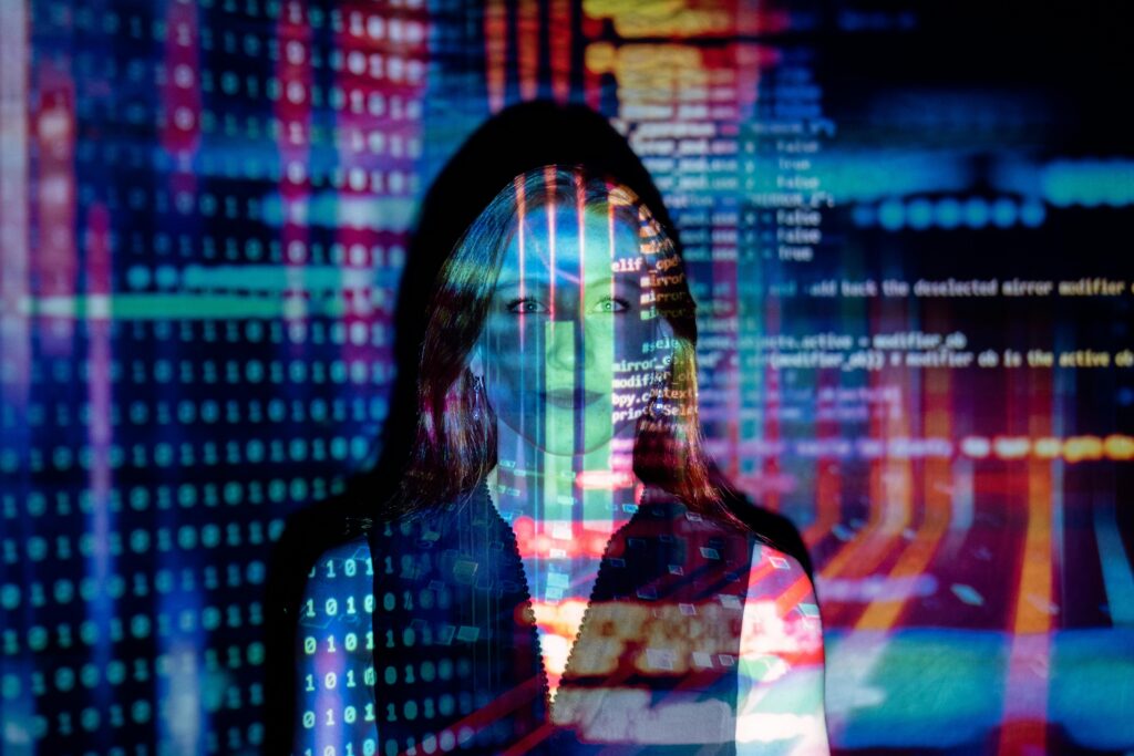 A person stands in front of a colorful backdrop filled with projected programming code, glowing binary numbers, and abstract digital patterns that light up their face and silhouette.